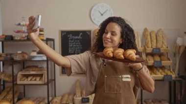 Kıvırcık saçlı bir kadın pastanede kruvasan tutarken arka planda raflarda ekmek sergilenirken selfie çekiyor.
