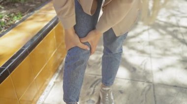 Gündüz kıyafetli genç bir kadın güneşli bir günde dışarıda dikilirken dizini acı içinde tutuyor..