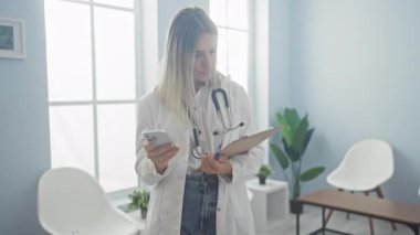 Beyaz önlüklü genç sarışın bir doktor akıllı telefon kullanıyor ve elinde parlak bir hastane bekleme odasında pano tutuyor..