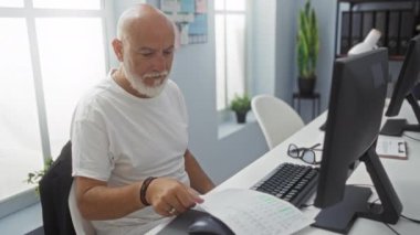 Üst düzey bir adam ofis ortamında evrak okurken, bir bilgisayarın yanında gözlüğünü takıyor..