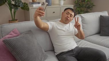 Genç Asyalı bir adam oturma odasında selfie çekiyor, rahat gri bir koltukta otururken huzur ve baş parmak hareketi yapıyor..