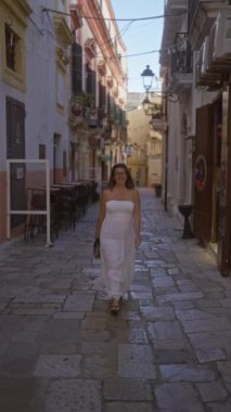 Beyaz elbiseli genç bir İspanyol kadın Puglia, İtalya ve Avrupa 'nın büyüleyici, dar caddelerinde yürürken gülümsüyor. Çevresinde canlı binalar ve açık hava oturma yerleri var..