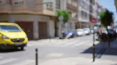 Defocused urban street with blurred yellow van and vehicles parked along the side under bright sunlight