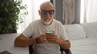 Gri saçlı, sakallı bir adam elinde kahve fincanı, elinde güneş gözlüğü ve beyaz gömlekle oturma odasında güneşli perdelerle oturuyor.