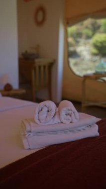 Yuvarlak havlular çadırın içinde düzgünce yapılmış bir yatağa yerleştirilmiş, arka planda sakin bir doğa manzarası olan, konforlu bir kamp macerasına işaret ediyor..