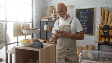 Beyaz saçlı ve sakallı orta yaşlı bir adam arka planda ekmek rafları ve hamur işleri olan bir fırında akıllı telefon kullanıyor..