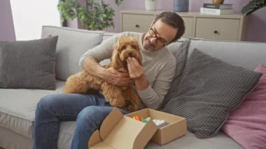 Orta yaşlı bir adam evde fino köpeğiyle oynuyor, rahat oturma odasında kutusunu açıyor.