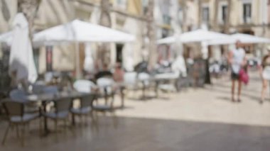 Güneşli bir günde odak noktasından çıkmış bir erkek ve kadının olduğu açık hava kafe sahnesinde masa sandalyeleri şemsiyeler ve bulanık bir arka plan görüntüsü var.