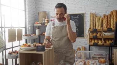 Fırında önlük giymiş orta yaşlı bir adam telefonda konuşuyor ve ekmek ve hamur işleriyle çevrili bir deftere yazı yazıyor.