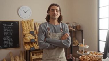 Kolları çapraz duran genç adam ekmek ve pasta vitrinleriyle çevrili, gri bir gömlek ve önlük giymiş, arka planda görünen saat ve fiyat tahtası