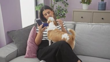 Genç bir İspanyol kadın oturma odasında oturuyor, elinde kredi kartı ve cep telefonu var, Chihuahua köpeğiyle iletişim kuruyor..