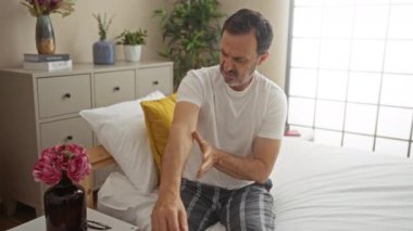 Yatak odasında kollarını ovuşturan orta yaşlı bir adam komodindeki çiçeklerle yatakta rahatsız görünüyor.