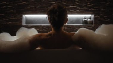 Rahat bir kadın loş, modern bir banyo ortamında lüks bir köpük banyosunun tadını çıkarır..