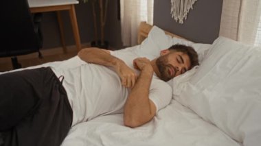 Sakallı genç bir adam uyanıyor ve sabah yatak odasında uzanıyor.