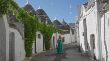 Yeşil elbiseli genç İspanyol kadın açık mavi gökyüzünün altında, ikonik trulli evler ve canlı yeşilliklerle çevrili, İtalya 'nın büyüleyici eski Alberobello kasabasında yürüyor..