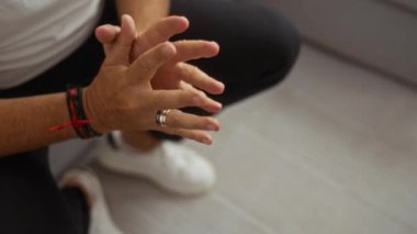 Oturma odasında elleri birbirine kenetlenmiş, yüzükle bilezik takmış siyah pantolon ve beyaz ayakkabılar giymiş bir adam.