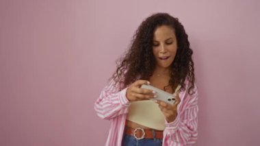 Kıvırcık saçlı, akıllı telefon kullanan, gülümseyen ve nişanlı genç bir kadın, izole edilmiş pembe arka plan duvarına karşı..