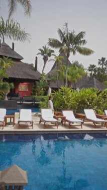 Bali 'de sakin bir tatil köyünün yanında genç bir kadın yürüyor. Güverte sandalyeleri ve tropikal bitki örtüsüyle çevrili..