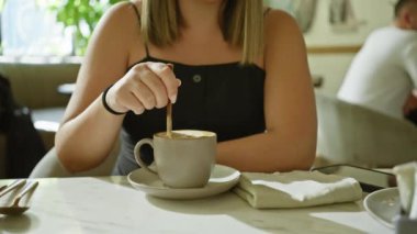 Yetişkin bir kadın, kafeterya masasında kahve içmekten hoşlanır. İçkisini evin içinde düşünceli bir şekilde karıştırır.