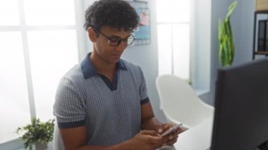 Zeki bir telefonla çalışan genç adam aydınlık, modern bir ofiste gülümsüyor.