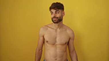 Gömleksiz genç İspanyol adam ayakta ve parmak ve elle işaret ediyor, izole edilmiş sarı arka planda mutlu bir şekilde gülümsüyor.