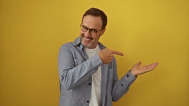 Orta yaşlı İspanyol bir adam, gözlük takıyor, ayakta duruyor ve el sallıyor, gülümsüyor ve izole edilmiş sarı arka planı işaret ediyor.