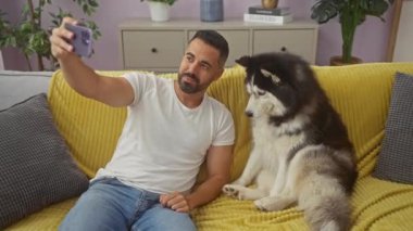 Gülümseyen İspanyol adam rahat oturma odasında köpeğiyle selfie çekiyor.