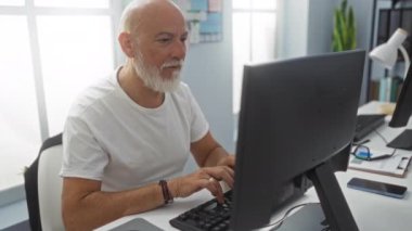Gri saçlı ve sakallı yaşlı bir adam bilgisayarlı bir ofis masasında çalışırken boynu ağrıyan parlak doğal ışıkla modern bir çalışma alanını gözler önüne seriyor..