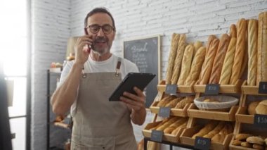Fırında orta yaşlı bir adam, arka planda ekmek dolu raflarla telefon görüşmesi yaparken elektronik tablet kullanıyor.