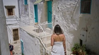 Beyaz elbiseli genç bir kadın, Ostuni, Puglia, İtalya 'nın dar sokaklarında yürüyor. Beyaz boyalı büyüleyici binalar turkuaz kapılar ve pencerelerle vurgulanıyor..
