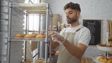 Genç İspanyol bir adam fırında çalışıyor. Rafta taze hamur işleri düzenliyor. Yüzünde odaklanmış bir ifade ve önlük var.