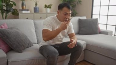 Evdeki bir salonda oturan genç adam, bir kanepede oturuyor ve akıllı telefonunu kullanıyor, modern kapalı bir ortamda bitkiler ve şık dekorlarla..