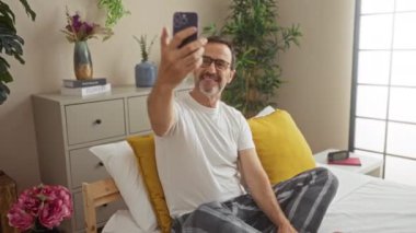 Adam rahat yatak odasında çiçek ve dekorasyonla selfie çekiyor, gözlük ve pijama giyiyor, sarı yastıklarla yatakta oturuyor.