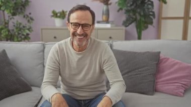 Gülümseyen, orta yaşlı, gözlüklü ve günlük kıyafetleri olan bir adam evdeki gri koltukta rahat bir şekilde oturuyor..