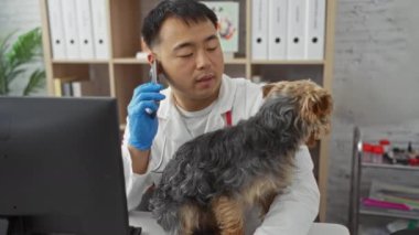 Veteriner kliniğindeki genç Çinli bir adam mavi eldivenli bir köpeği tutuyor ve profesyonel bir iç mekanda bilgisayar başında çalışırken telefonla konuşuyor..