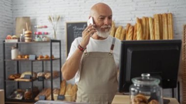 Kel kafalı, gri sakallı, fırının içinde önlük takan, telefonla konuşan ve çeşitli ekmek ve hamur işleriyle çevrili bir bilgisayarı işleten olgun bir adam..