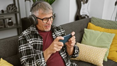 Kulaklıklı, neşeli, orta yaşlı bir adam rahat bir oturma odasında akıllı telefon kullanıyor. Bu da modern boş zamanların göstergesi..