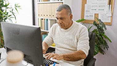 Orta yaşlı bir İspanyol, modern bir ofis ortamında bilgisayarının başında çalışmaya odaklanmış..