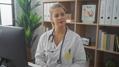 A blonde, professional woman in a white coat adorned with a stethoscope sits by a computer in a well-lit clinic office. clipart