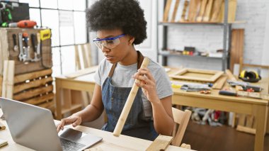 Marangozluk atölyesinde elinde odun tutarken dizüstü bilgisayar kullanan odaklı bir Afrikalı Amerikalı kadın..