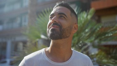 Yakışıklı İspanyol bir adam güneşli bir şehir ortamında düşünceli bir şekilde yukarı bakıyor, arka planda yeşil ve şehir binaları bulanık.