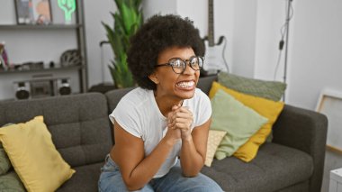 Heyecanlı, kıvırcık saçlı, gözlüklü, ev içinde gri bir koltukta oturan Afro-Amerikalı kadın..