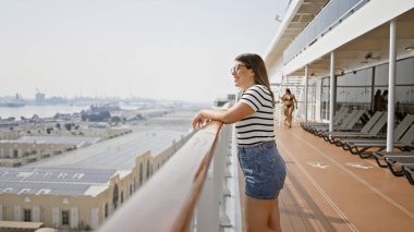 Smiling woman leaning on cruise ship railing overlooking port on sunny day clipart