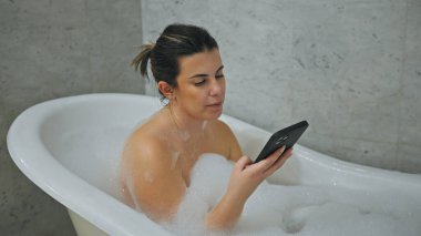 Genç yetişkin bir kadın evdeki köpük banyosunda dinleniyor, sakin bir banyoda akıllı telefonuna bakıyor..