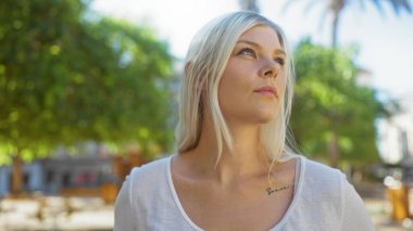 Young woman with blonde hair wearing a white shirt in an urban park during the day with trees and buildings in the background. clipart