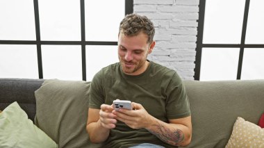 Sakallı, gülümseyen, genç İspanyol bir adam. Kapalı bir kanepede akıllı telefon kullanıyor..