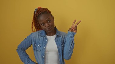 Kot pantolonlu genç Afrikalı kadın sarı arka planda izole edilmiş barış işareti yapıyor.
