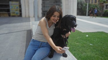 Genç bir İspanyol kadın şehir parkında güneşli bir günün tadını çıkarıyor, dışarıda bankta otururken mutlu siyah labradoruyla selfie çekiyor..