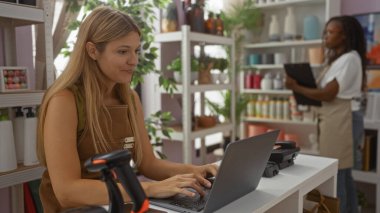 Ev dekorasyonu mağazasında dizüstü bilgisayarda çalışan bir kadın, arka planda raflar düzenleyen, çeşitli dekoratif eşyalar ve bitkilerle ev içi iş ortamını gösteren bir iş adamı.