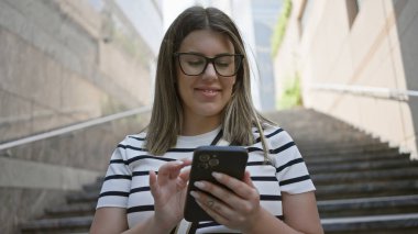 Arka planında modern mimari olan akıllı telefon kullanan gülümseyen kadın..